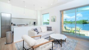 A modern kitchen with a stainless steel refrigerator, a white countertop, and a coffee machine. There is a white sofa with patterned cushions and a small table with chairs. A large window offers a view of a body of water and greenery. - Cayman Luxury Rentals at One Canal Point (Seven Mile Beach)
