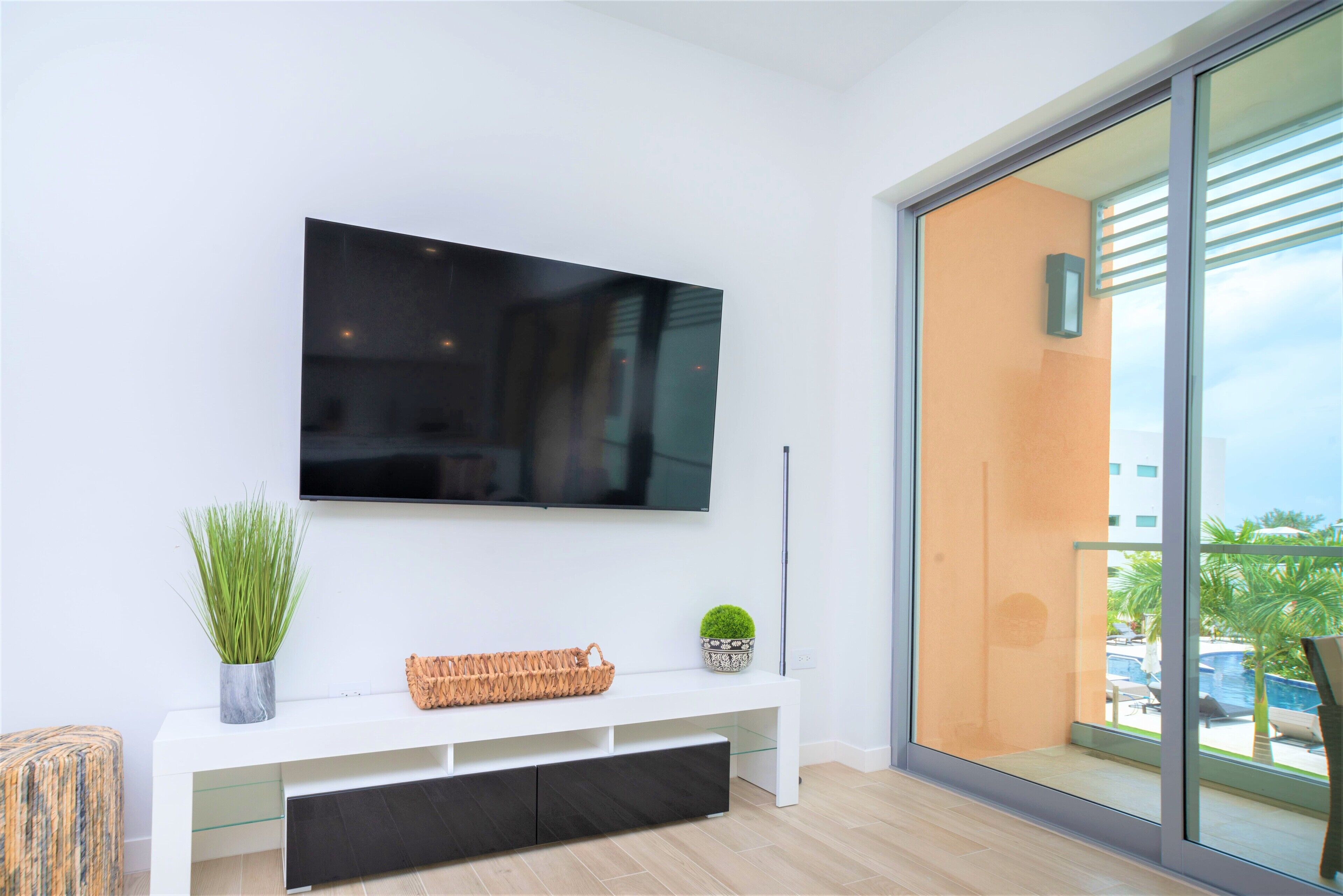 Een moderne woonkamer met een flatscreen televisie aan de muur, een witte console tafel met een mand, een kleine potplant en een schuifdeur naar een buitenruimte.
