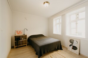 A bedroom with a bed, a wooden shelf with decorative items, a window with blinds, and a light fixture on the ceiling.