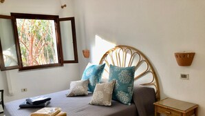 Quarto com cama, almofadas, mesa de cabeceira, janela e luminária na parede.
