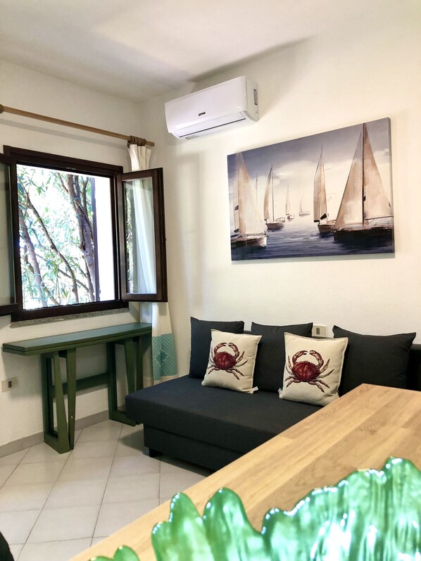 A room with a dark green sofa, two crab-themed pillows, a wooden table, and a painting of sailboats on the wall.