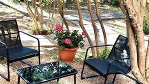 Uma área externa com assentos, incluindo quatro cadeiras de metal preto e uma mesa de vidro, rodeada por árvores e um vaso de plantas com flores cor-de-rosa.