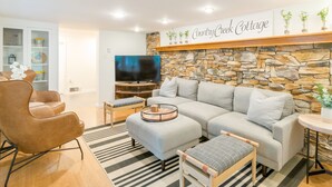 A modern living room with a stone wall, a television, a sofa, a coffee table, and a bookshelf. - PENN STATE Country Creek Cottage w/ Hot Tub - ON Spring Creek and 7 miles to PSU (Bellefonte)