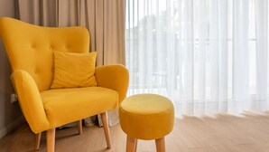 Uma poltrona amarela com um banquinho a combinar, num quarto com soalho de madeira clara e cortinas leves.
