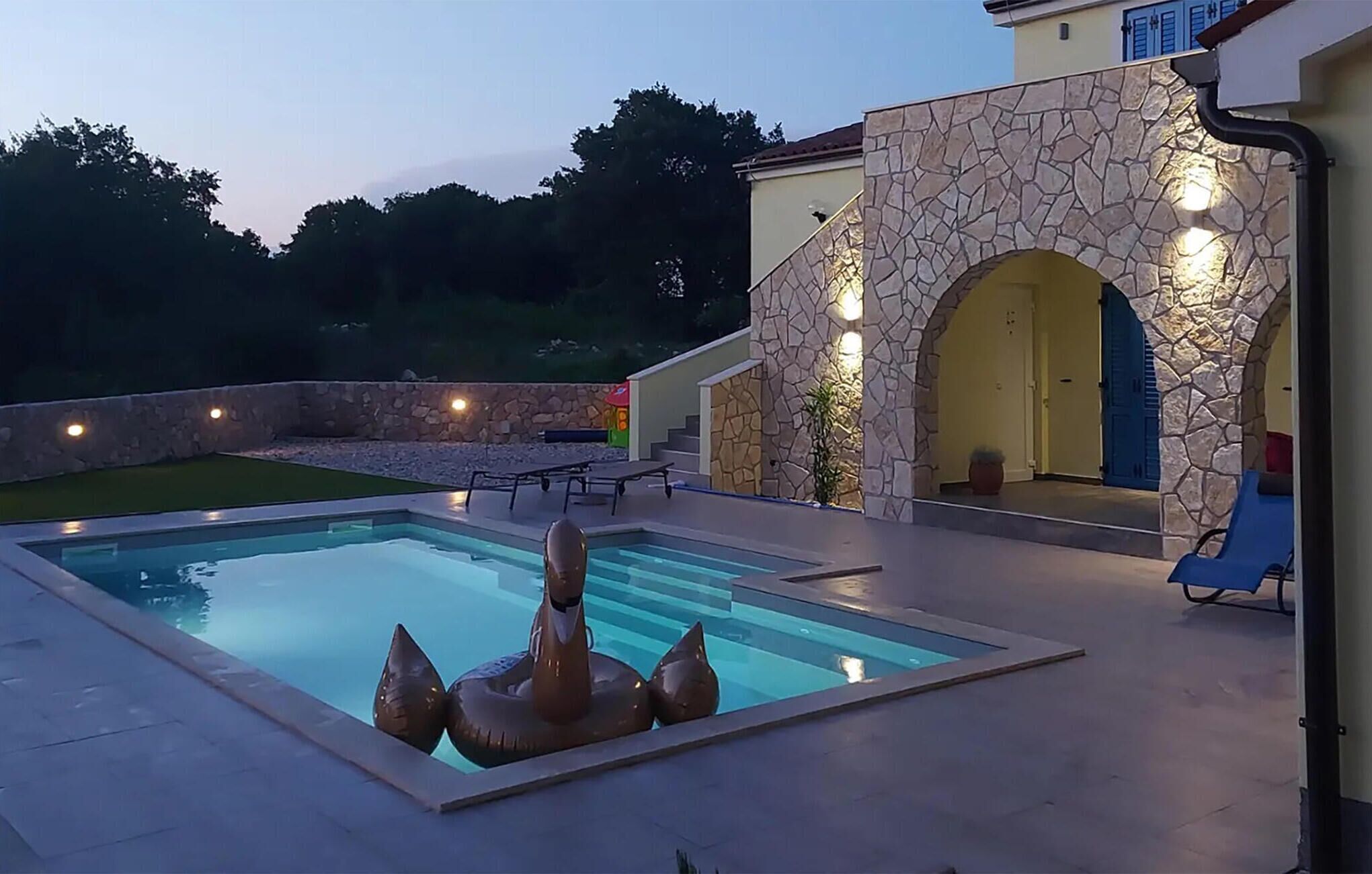 Una piscina con un muro de piedra y una puerta, rodeada por un patio con mobiliario de exterior.