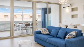 Ein modernes Wohnzimmer mit einem blauen Sofa, gemusterten Kissen, einer Glastür zu einem Essbereich im Freien und einer weißen Wand mit Einbaustrahlern.