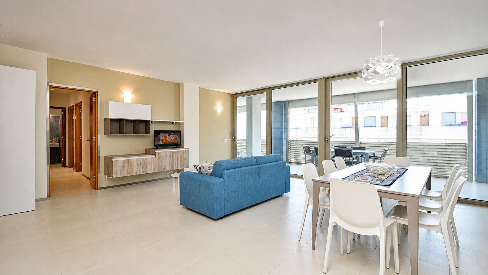 Ein modernes Wohnzimmer mit einem blauen Sofa, einem Essbereich mit Tisch und Stühlen, einer Küchenzeile mit Schränken und einem Fernseher, sowie großen Fenstern mit Blick.