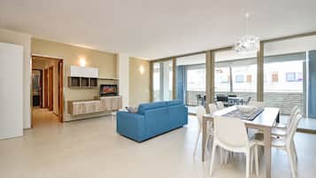 Ein modernes Wohnzimmer mit einem blauen Sofa, einem Essbereich mit Tisch und Stühlen, einer Küchenzeile mit Schränken und einem Fernseher, sowie großen Fenstern mit Blick.