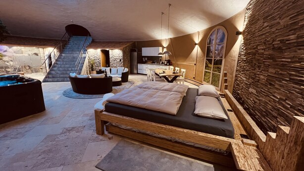 A spacious bedroom with a large bed, a stone wall, and a view of a mountainous landscape. - Wellness-Speicher vacation apartment with sauna, whirlpool, breakfast and more (Freital)