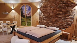 A modern bedroom with a wooden bed, a round bench, a dining table with chairs, and a large window with a mountain view. - Wellness-Speicher vacation apartment with sauna, whirlpool, breakfast and more (Freital)