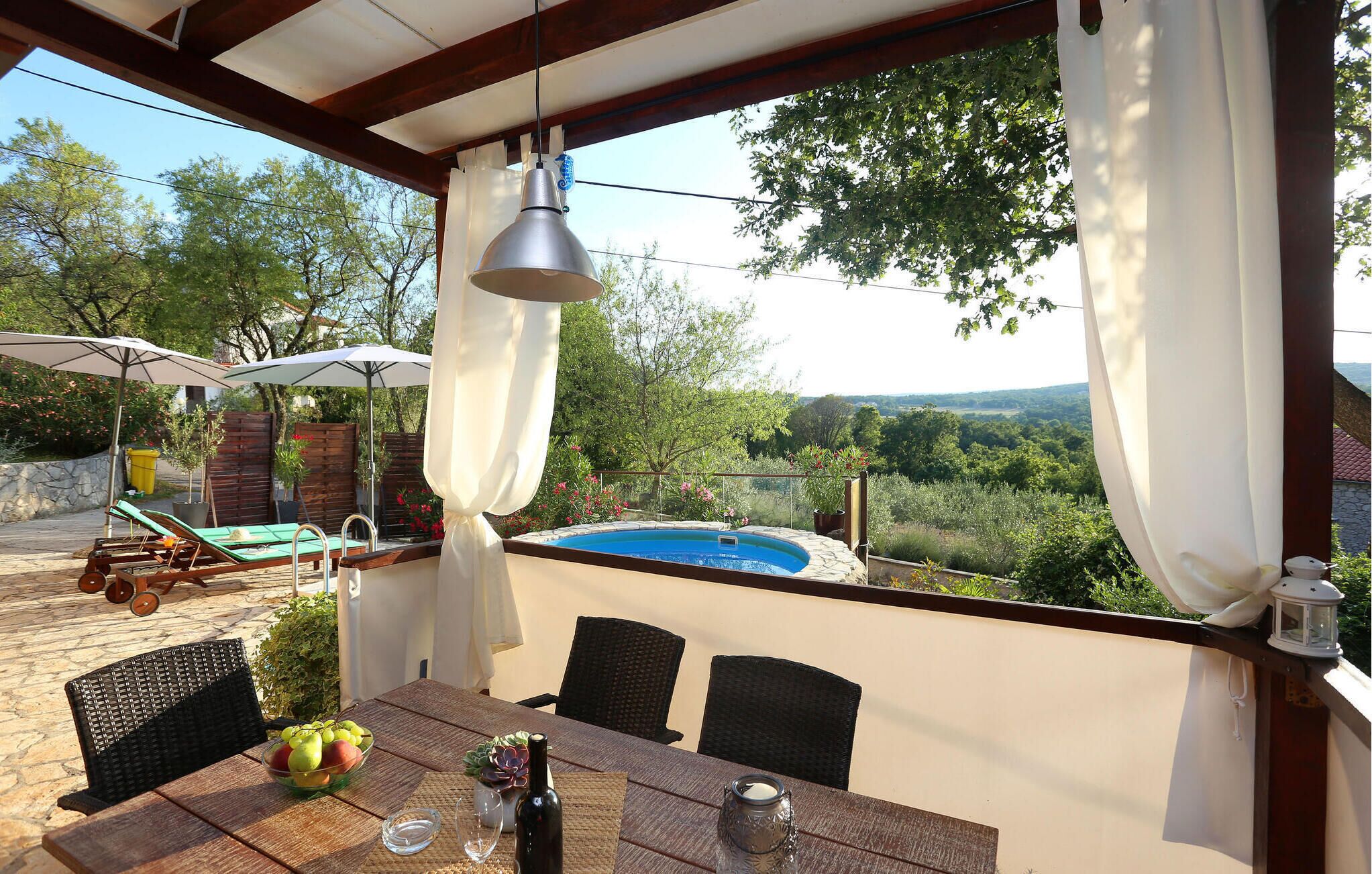 Un espacio al aire libre techado con piscina, sillas de descanso, una mesa con frutero y vistas del paisaje circundante.