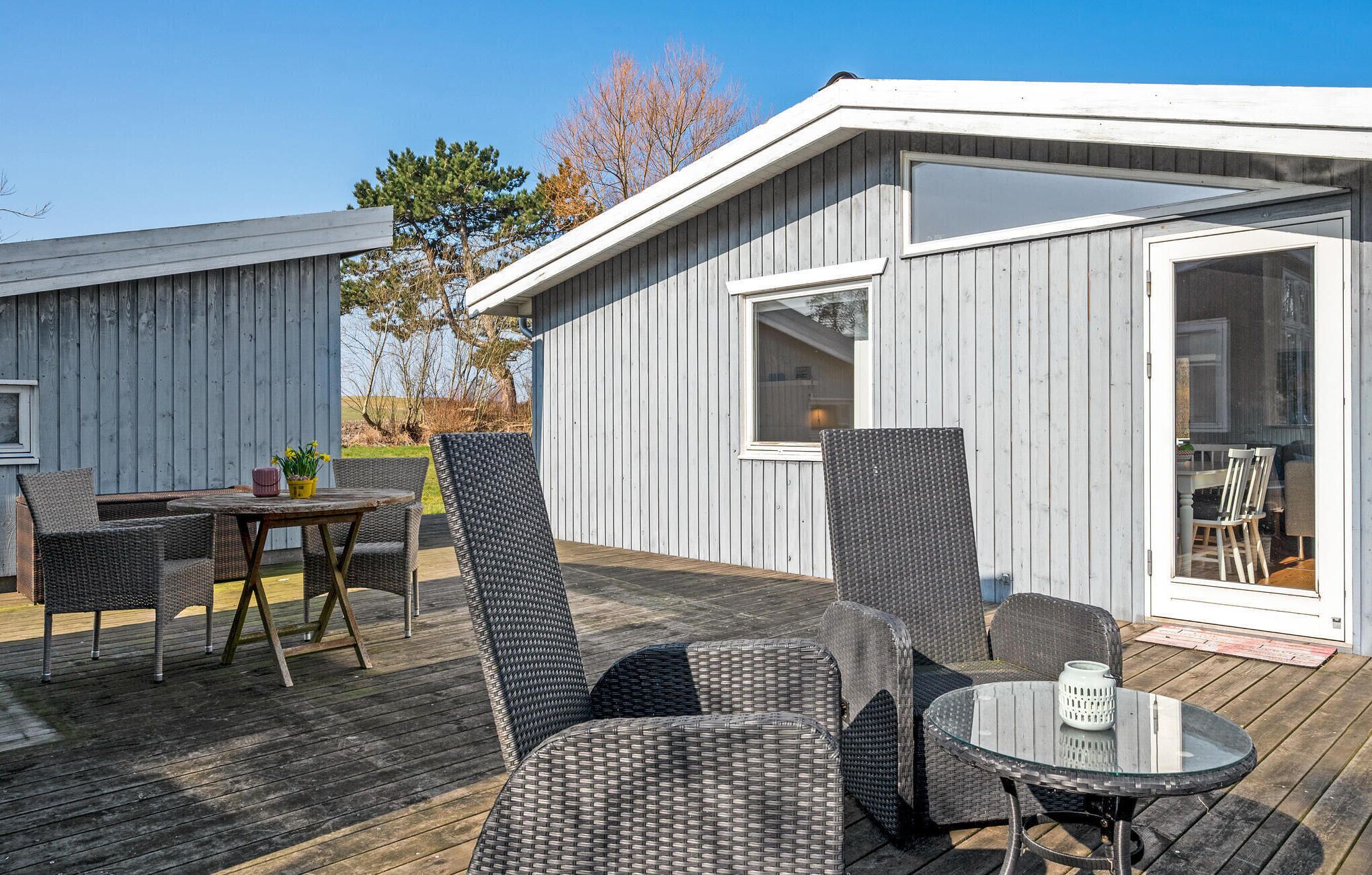 Een overdekt buitengebied met rieten meubels en een glazen tafel, naast een gebouw met een schuifpui.