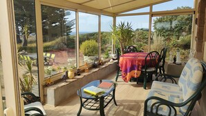 A sunlit patio with a table and chairs, a glass table, and a view of a garden and hills. - Maison Vue mer avec grand jardin sur la Côte de Granit Rose (Pleumeur-Bodou)