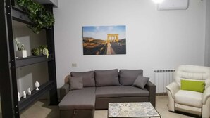 A living room with a grey sofa, a white armchair with a green cushion, a coffee table with a patterned top, a wall-mounted air conditioner, and a shelf with plants and decorative items.