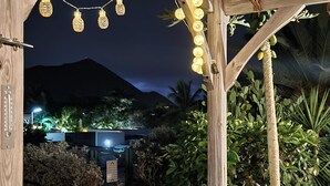 Una estructura de pérgola de madera con luces colgantes, rodeada de exuberante vegetación y una montaña al fondo.