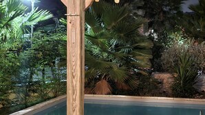 Terraza de madera con piscina, rodeada de plantas tropicales y palmeras, iluminada con luces colgantes.