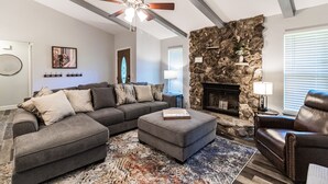 A living room with a sectional sofa, ottoman, fireplace, stone accent wall, ceiling fan, and a rug.
