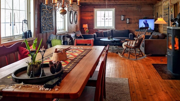A cozy cabin interior with a wooden dining table, a sofa, a rocking chair, and a fireplace.