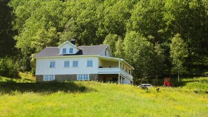 A two-story house with a balcony, surrounded by a grassy field and dense trees.
