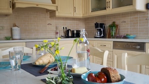 Une cuisine avec des meubles de couleur beige, un plan de travail en marbre et un lave-vaisselle. On y trouve une table avec une nappe bleue, une bouteille en verre et un saladier rempli de tomates.
