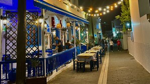 Une scène de rue nocturne avec un restaurant doté d’une terrasse, de guirlandes lumineuses et d’une décoration aux couleurs bleu et blanc.