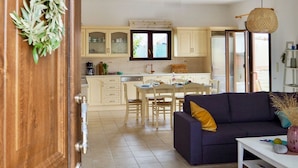 Une cuisine moderne avec des meubles blancs, un coin repas avec une table et des chaises, et un salon avec un canapé et un luminaire suspendu.