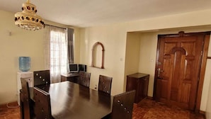 A dining area with a large wooden table, six chairs, a chandelier, a water dispenser, and a wooden door. - Spacious 3 bedroom with pool (Nairobi)