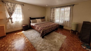 A bedroom with a wooden floor, a bed with a patterned bedspread, two nightstands, a dresser, and a large window with curtains. - Spacious 3 bedroom with pool (Nairobi)