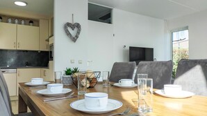 Comedor con una mesa de madera dispuesta para cuatro, platos, tazas y cubiertos blancos. Una decoración con forma de corazón cuelga de la pared, y un televisor está montado sobre la mesa.