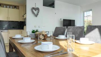 Een eethoek met een houten tafel gedekt voor vier personen, witte borden, bekers en bestek. Een hartvormige decoratie hangt aan de muur, en boven de tafel is een televisie bevestigd.