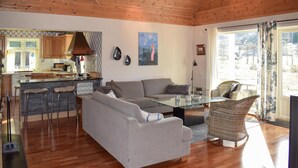 Ein modernes Wohnzimmer mit einem grauen Sofa, einem Glastisch und Korbstühlen. Im Hintergrund ist eine Küche mit Holzausbau und einem Fenster erkennbar.