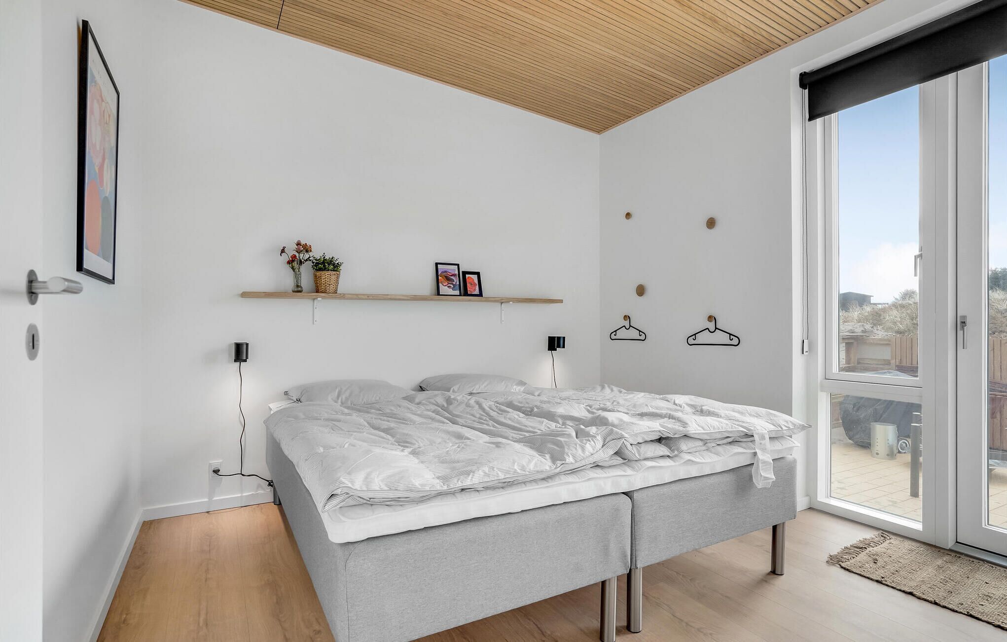 Quarto moderno com cama, teto de madeira, paredes brancas e uma janela grande.