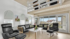 Ein modernes Wohnzimmer mit einer schwarzen Ledersofa, einem Holztisch und schwarzen Ledersesseln. Große Fenster bieten Blick auf Häuser und einen Strand.