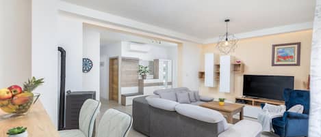 En moderne stue med en grå hjørnesofa, en blå lænestol, et fjernsyn og et spiseområde med bord og stole.