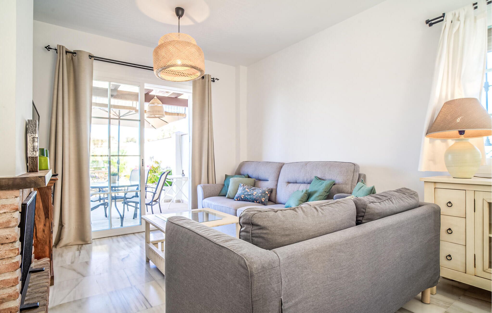 Un salon avec un canapé gris, une table et une lampe. Il y a une fenêtre avec des rideaux et une vue sur l’extérieur.