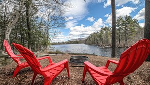 Zwei rote Adirondack-Stühle blicken auf einen See, dazwischen ein Feuerkorb, umgeben von Bäumen und einem klaren Himmel.