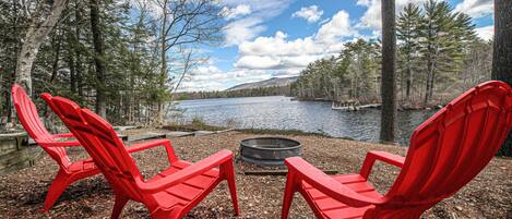 Two red Adirondack chairs facing a lake with a fire pit in between, surrounded by trees and a clear sky.