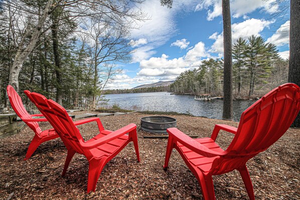 Two red Adirondack chairs facing a lake with a fire pit in between, surrounded by trees and a clear sky.