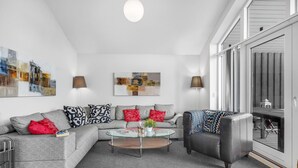 Een moderne woonkamer met een grijze modulaire bank, een zwarte fauteuil en een glazen salontafel. Aan de muren hangen abstracte schilderijen en er is een klein balkon zichtbaar door de glazen deur.