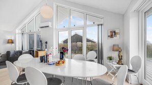 Een moderne eetruimte met een grote ronde tafel, witte stoelen en uitzicht op een houten gebouw door grote ramen.