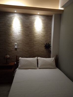A bedroom with a stone-textured wall, a bed with white bedding, and a nightstand with a lamp.