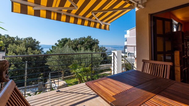A balcony with a wooden table and chairs, a striped awning, and a view of the sea and trees. - Beautiful home in Marusici (Omis) with WiFi (Marusici (Omis))