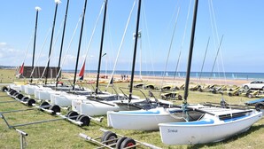 Uma fileira de barcos à vela atracados em uma área gramada, com uma praia e oceano ao fundo.