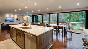 A modern kitchen with a large island, wooden cabinets, and a view of a deck and trees.