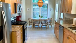 A modern kitchen with wooden cabinets, stainless steel appliances, a dining area with a table and chairs, and a large window providing natural light.