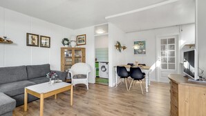 Ein modernes Wohnzimmer mit grauem Sofa, einem weißen Couchtisch, einem Korbstuhl und einem Essbereich mit Tisch und Stühlen. Im Hintergrund steht eine Waschmaschine, und ein Fernseher auf einer Holzkommode.