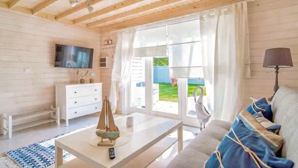 Un salon moderne avec un téléviseur, un canapé blanc agrémenté de coussins bleus, une table basse et une vue sur un jardin grâce à de grandes fenêtres.