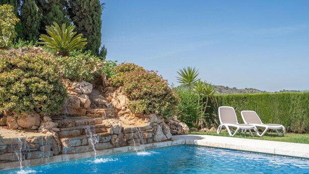 A swimming pool with a stone waterfall feature, surrounded by lush greenery and two white lounge chairs. - 1 bedroom cozy apartment in Mondron (Mondron)