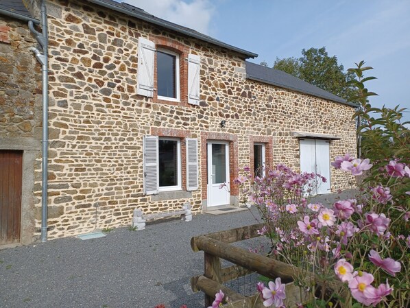 Un bâtiment en pierre avec trois fenêtres, une clôture en bois et des fleurs roses au premier plan.