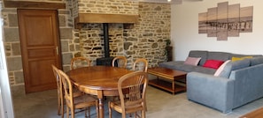 Un salon chaleureux avec un mur en pierre, une table à manger en bois avec des chaises, un canapé gris agrémenté de coussins colorés et un grand tableau accroché au mur.